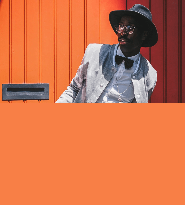 Modelo mascuilno utilizando óculos escuros e vestindo terno na cor branco com chapéu preto, em um fundo laranja.